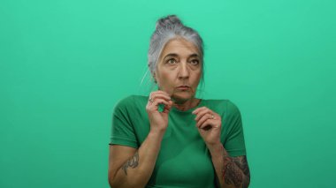 Yeşil tişörtlü yaşlı, gri saçlı kadın izole edilmiş yeşil bir arka plan duvarına karşı poz veriyor, anlamlı jestler sergiliyor..