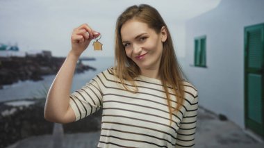 Gülümseyen kadın, gün ışığında sahil kenarında küçük ahşap ev anahtarlığını nazikçe tutuyor; ev sahipliği gururu.