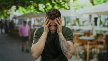 Dövmeli genç adam güneşli bir şehir terasında stresli görünüyor. Etrafı hareketli şehir hayatı ile çevrili. İç duygular ve canlı çevre arasındaki zıtlığı yakalıyor..