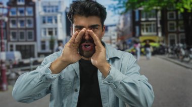 Kot ceketli adam şehir caddesinde bulanık binalar ve geçen bisikletlerle bağırırken ellerini ağızlarına uzatıyor; acil çağrı.