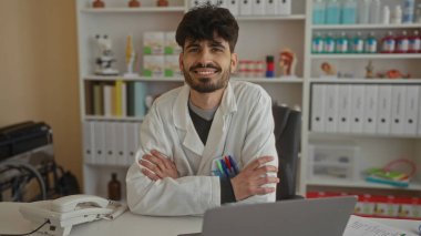 Klinikte gülümseyen genç bir adam arka planda raflar ve tıbbi ekipmanlarla profesyonel bir çevreyi ön plana çıkarıyor..