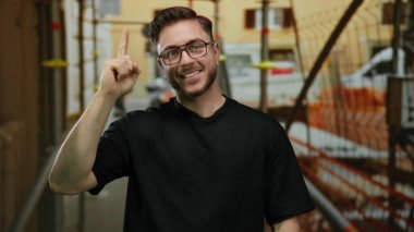 Gündüz vakti arka planda iskelesi olan bir inşaat alanında gözlük takan sakallı genç bir adam..