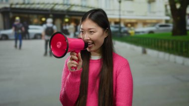 Pembe kazaklı uzun saçlı kadın şehir sokaklarında megafonla konuşuyor, şehir hayatı ve aktiviteleriyle çevrili, açık hava ortamında kendine güvenen iletişimi somutlaştırıyor..