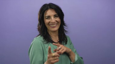 Mor stüdyoda yeşil bluz giyen, gülümseyen ve kamerayı işaret eden İspanyol kadın..