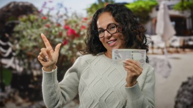 Dışarıda gülümseyen bir kadın elinde Dirham banknotuyla heyecanla bir şehir caddesini işaret ediyor..