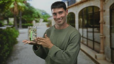 Latin asıllı genç bir adam, yeşil parkta bir elinde şeffaf dolar kavanozu tutarken, tasarruf iyimserliği....