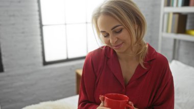 Kadın yatak odasında huzurlu bir sabah geçirir, rahat bir evde sıcak bir içkinin tadını çıkarır, kırmızı bir bluz giyer, sükunet ve rahatlama anı yakalar..