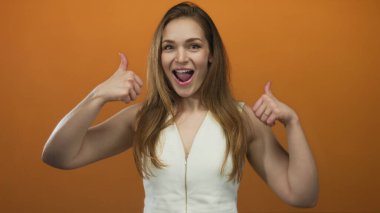 Bir stüdyo ortamında canlı turuncu bir arkaplan karşısında başparmak hareketi yapan ve gülümseyen bir kadın..
