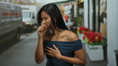 Genç bir kadın şehir caddesinde öksürüyor, rahatsızlığını dile getiriyor, dalgalı saçlı mavi bir elbise giyiyor, çevresi çiçeklerle çevrili..