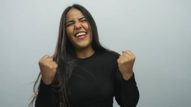 Beyaz bir arkaplan karşısında yumruklarını kaldırarak kutlama yapan bir kadın stüdyo ortamında zafer dolu bir pozla neşe ve başarısını ifade ediyor..