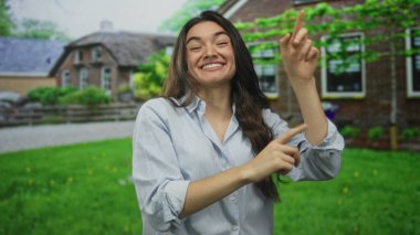 Parlak gülüşlü bir kadın yeşil çimenlikteki sokakta tuğla evin girişini işaret ediyor. İyimserlik..