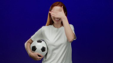 Futbol topunu tutan kadın canlı ve enerjik bir spor teması içinde kırmızı saçlarını ve günlük beyaz gömleğini sergileyerek neşeli bir şekilde mavi arka plana gülümsüyor..