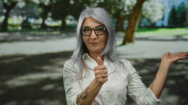 Gri saçlı, gözlüklü, baş parmağını kaldıran ve parkta neşeli bir ifade ve kolları dövmeli bir şey sunan son sınıf öğrencisi kadın..