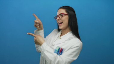 Laboratuvar önlüklü, kırmızı gözlüklü bir kadın, canlı mavi bir duvarın karşısında duruyor, şaşkınlık ve coşkuyu dile getiriyor, bir keşif hissi veriyor..