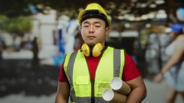 Elinde emniyet yeleği ve miğfer takan, hareketli bir şehir caddesinde duran, şehir mimarisini ve inşaatını Asya bağlamında temsil eden kendine güvenen genç bir adam..