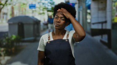 Önlük giymiş bir kadın, şehir caddesindeki açık alanda düşünceli bir şekilde duruyor..