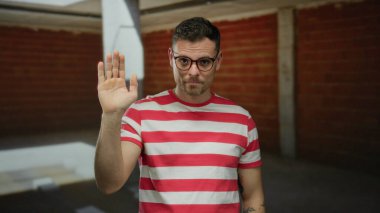 Gözlüklü genç bir adam inşaat alanında el sallıyor, kırmızı çizgili bir gömlek giyiyor, kendine güveni ve günlük davranışları var..