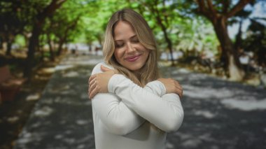 Gözleri kapalı sarışın bir kadın ve yeşil parkta nazik bir gülümseme ile omuzlarına sarılıyor..