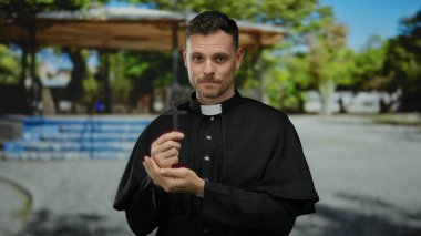 Siyah cüppeli Katolik bir rahip elinde bir haç, açık hava parkı ortamında, güneş ışığı altında huzur ve yansıma simgeliyor..