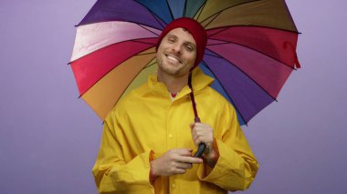 Renkli şemsiyelerin altında gülümseyen genç adam mor arka planda sarı yağmurluk giyiyor..