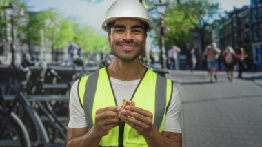 Bisiklet raflarının önündeki kalabalık şehir caddesinde gözleme yiyen adam sarı şapka ve sarı güvenlik yeleği takıyor; memnuniyeti kırın.