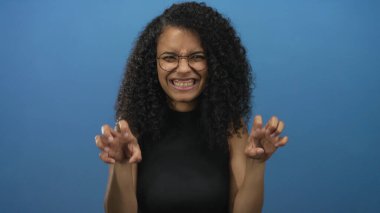 Mavi stüdyoda pençe jesti ve gülümseyen yüzüyle gözlük takan bir kadın; neşe dolu bir eğlence.