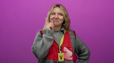 Kırmızı yelek ve gri süveter giyen genç gönüllü kadın, elleri çenesinde, pembe arka planda izole edilmiş bir şekilde gülümsüyor, neşeli bir atmosferde bir fikir öneriyor..