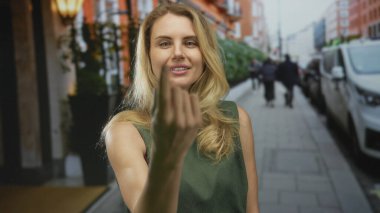 Şehir caddesinde elini kolunu sallayarak gezen, sarı saçlı, yeşil elbiseli, canlı bir şehir ortamında, park halindeki arabaların ve bulanık yayaların arasında davetkar bir şekilde el kol gezen bir kadın..