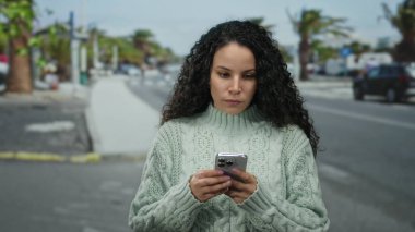Sokakta akıllı telefon kullanan bir kadın kıvırcık saçlı bir kazak giyiyor. Odaklanmış ifade, gündüz vakti şehir ortamında teknolojiyi gösteriyor.