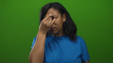 Mavi gömlek giyen Afrikalı Amerikalı kadın yeşil stüdyo duvarına kapalı gözleri olan burnunu çimdikliyor..