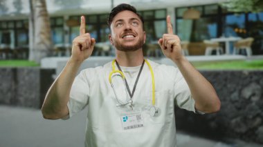 Şehir ortamında sakallı ve steteskoplu genç bir adam şehrin caddesinde dikilirken iki eliyle yukarı doğru bakıyor..