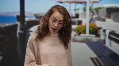 Gözlüklü bir kadın, deniz kenarında güneşli bir günde etrafı manzarayla çevrili, rahat bir kazak giymiş, kumsal arkaplanı ve canlı çiçekleri olan bir kadın..