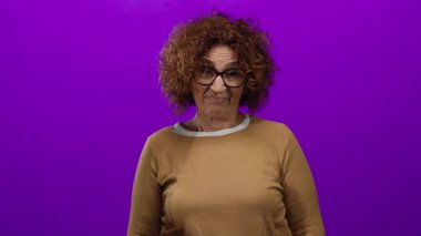 Elleriyle ezik bir jest yapan, gözlüklü ve kahverengi bir süveter giyen bir kadın, canlı mor bir arka plana karşı, duygularını ifade eden orta yaşlı, Hispanik bir kadına yer veriyor..