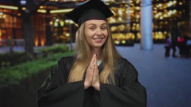 Mezuniyet kıyafeti giymiş genç bir kadın, bir şehir caddesinde, elleri minnettarlık jesti içinde, açık havada neşe ve başarıyı ifade ediyor..