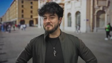 Yoğun bir caddede sakallı genç bir adam güvenli bir şekilde gülümsüyor tarihi binalar ve arka planda yürüyen insanlar kentsel yaşam tarzını ve kültürünü gözler önüne seriyor..