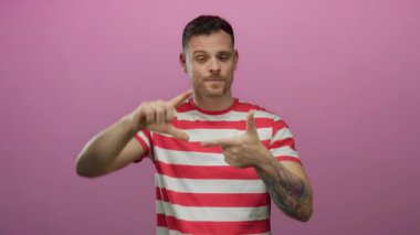 Çizgili gömlekli genç adam pembe arka planda poz veriyor. El çerçevesi hareketi yapıyor. Yaratıcı ifade ve tarz gösteriyor..