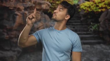 Latin kökenli genç adam, bulanık, kayalık arka plana karşı baş parmak hareketi yapıyor..