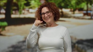 Orta yaşlı İspanyol bir kadın açık hava parkında kameraya bakıp gülümseyen, doğal ışık altında mutlu bir konuşma öneren bir jest yapıyor..