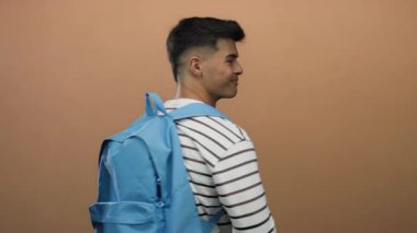 Sırt çantalı genç adam gülümsüyor ve izole bir kahverengi duvara başparmak işareti veriyor. Neşeli ve kendinden emin bir tavır sergiliyor..