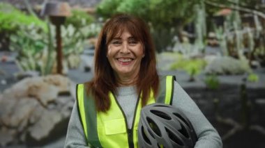 Parktaki son sınıf öğrencisi İspanyol kadın elinde bisiklet kaskı tutarken parlak bir şekilde gülümseyen bir güvenlik yeleği giyiyor. Etrafı yemyeşil ve sakin bir açık hava koşullarıyla çevrili..