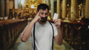 Katolik kilisesinde kredi kartı gösteren sakallı İspanyol bir adam geleneksel dini bir ortamda modern mali işlemleri vurguluyor..