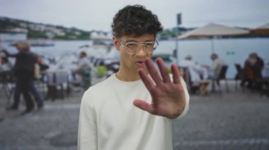 Gözlüklü ve beyaz tişörtlü İspanyol genç adam restoran terasında durmak için elini uzatıyor..