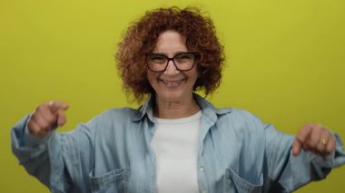 Kıvırcık esmer saçlı, gözlüklü ve mavi gömlekli bir kadın izole edilmiş sarı bir arka planın önünde coşkuyla el kol hareketi yapıyor..