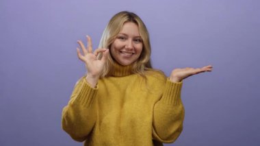 Mor arka plan üzerinde sarı kazak giyen kadın, gülümseyip avucunu gösterirken, pozitifliğini vurgularken ve neşeli tavrını sergilerken iyi işaret ediyor..