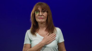 Gözlüklü yaşlı İspanyol kadın, beyaz tişört giyiyor, mavi duvara yaslanmış, el ele tutuşmuş, yemin ediyor ya da söz veriyor, izole edilmiş bir arka plan..