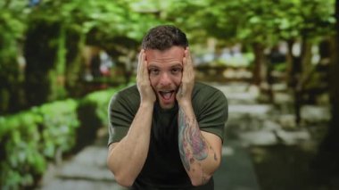 Dövmeleri ve kısa saçı olan genç bir adam, yemyeşil bir parkta, benekli güneş ışığı altında stresi ifade ediyor..