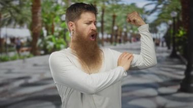 Beyaz gömlekli adam, yemyeşil yemyeşil parkta, palmiye astarlı yürüyüş yolunda pazılarını esnetiyor..