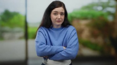 Mavi kazaklı, kumral saçlı bir kadın yeşil parkta dikiliyor bulanık arka plana karşı anlamlı bir jest sergiliyor..