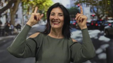 Orta yaşlı İspanyol kadın, güneşli şehir caddesinde parmaklarını yukarı kaldırarak gülümsüyor; umut iyimserliği.