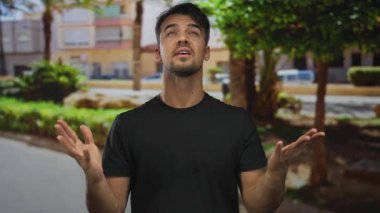 Genç İspanyol bir adam açık havada, yemyeşil bir parkın arka planında, arka planda konut binaları bulunan hareketli bir jest yapıyor..